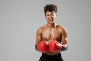 A young man boxer, displaying a confident smile, stands with his red boxing gloves raised. The studio setting features a neutral background, emphasizing his physique and dedication to the sport