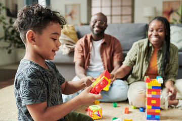 Obraz premium Side view of adorable little boy in casual attire sitting on the floor in front of his parents and playing constructor at leisure