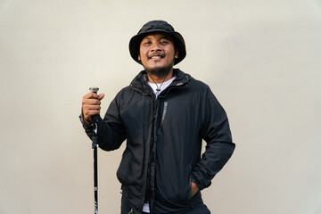 Photo of Asian man wearing all black adventure outfit. Half body portrait of an adult adventurer posing wearing a jacket, boonie hat and trekking poles isolated on a beige background