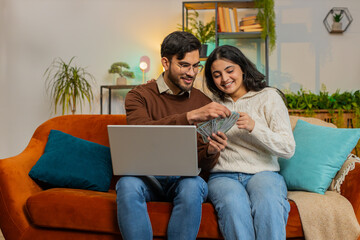 Happy young Arabian couple counting money and booking tickets for holiday vacation sits on sofa in living room at home. Diverse family holding banknotes planning online investment budget home finance