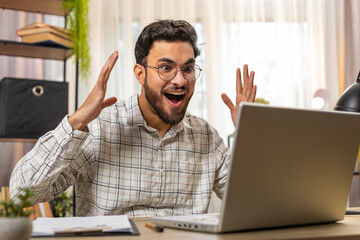 Cheerful amazed Indian businessman working on laptop shocked by sudden victory, game winning lottery goal achievement, good career news. Excited Arabian guy freelancer clenching fists at home office.