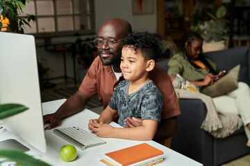 Young African American man in casual attire embracing his cute little son while both sitting by desk in front of computer monitor
