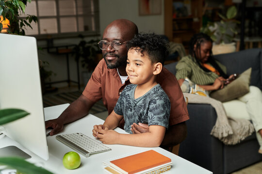 Young African American man in casual attire embracing his cute little son while both sitting by desk in front of computer monitor