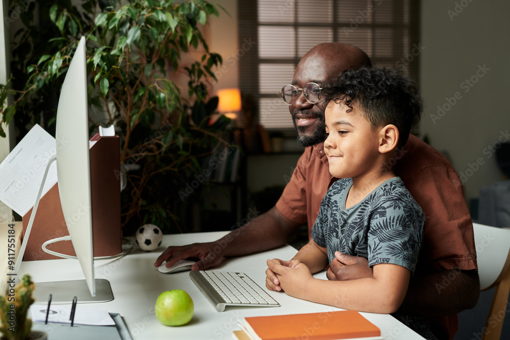 Poster happy cute little boy sitting by desk next to his father and both looking at desktop computer monito