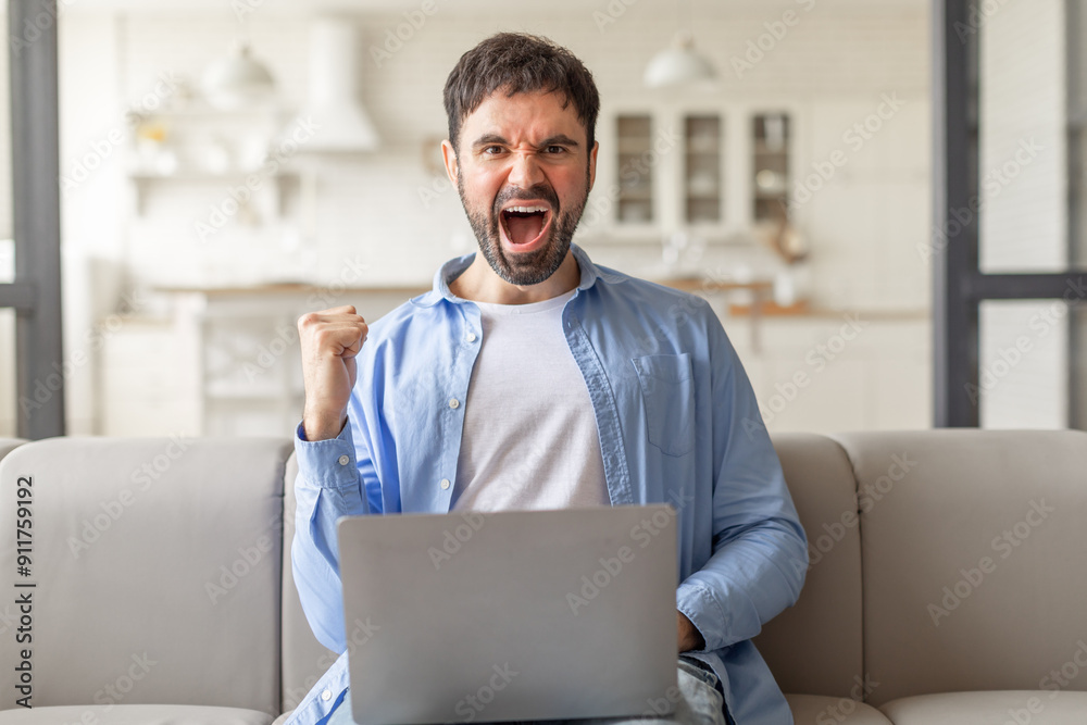 Poster a man with a beard sits on a beige couch in a living room and excitedly celebrates with a raised fis