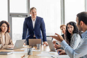 A group of business professionals are gathered around a table, discussing ideas. One man is standing, looking at the group while another man gestures, engaged in the conversation