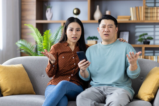 Asian couple sitting on sofa at home, both looking confused while holding smartphone. Concept of frustration, technology issue, misunderstanding, home lifestyle.