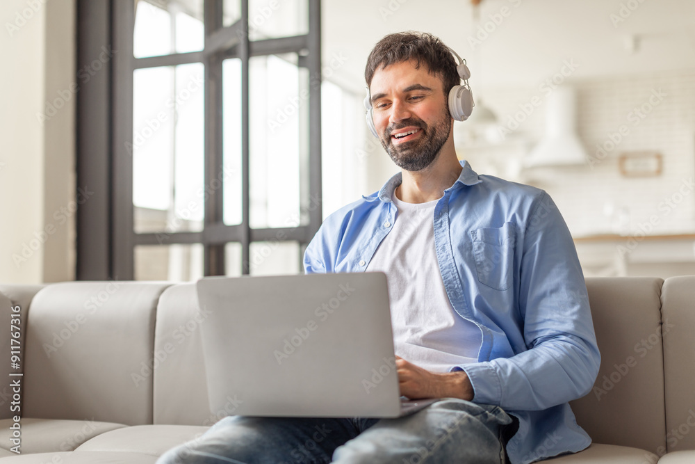 Poster a man sits on a white couch in a modern living room, wearing headphones and smiling while using a la
