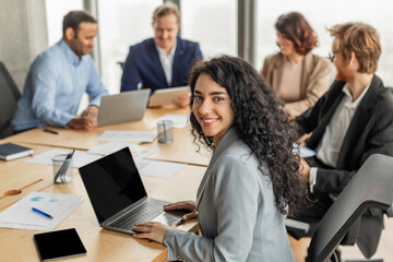 A young woman with curly dark hair smiles at the camera while sitting at a conference table with three other people. She is working on a laptop and the others are using tablets and computers.