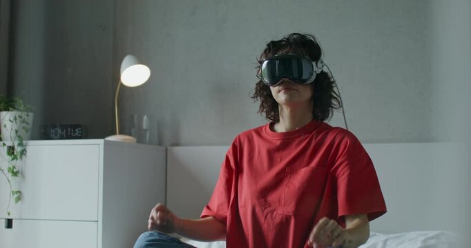 A woman in casual clothes and wearing a virtual reality headset moves her arms while sitting on a bed at home. Daylight