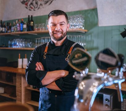 Portrait of happy smiling bearded barman dressed in a black uniform with an apron at bar counter with draught beer taps. Successful people, hard work, consumer cafes and restaurants industry concept.