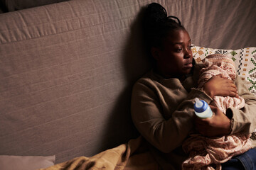 Miserable young woman sitting on couch at home and embracing newborn baby wrapped into cloth after...
