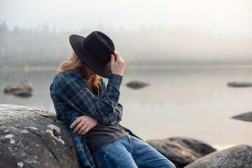 young man with long hair, cowboy hat, flannel shirt, and jeans, sitting by mountain lake