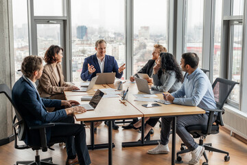 A group of business professionals are gathered around a large conference table in a modern office, engaging in a discussion. Laptops and documents are spread out on the table