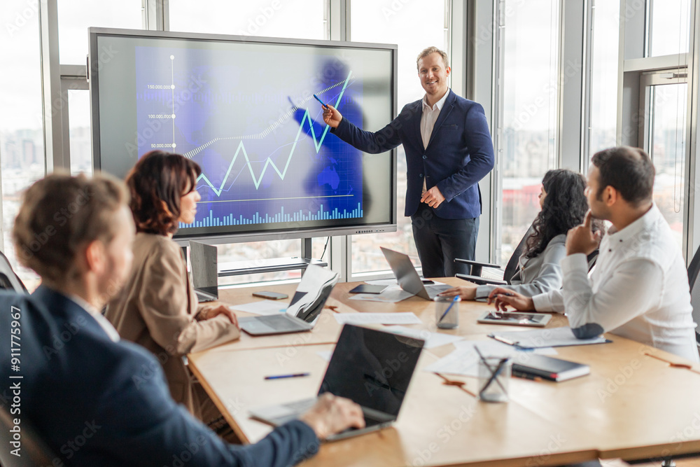 Poster A business professional stands near a large digital screen showing an upward trending line graph. The presenter gestures towards the screen as they explain the chart to a group of colleagues