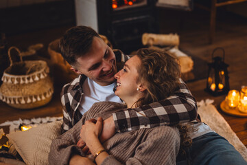 Happy young couple spending lazy weekend at home, embracing, kissing, enjoying near fireplace in the countryside cottage house, feeling relaxed and calm. Cozy home atmosphere, simple domestic life