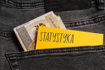Yellow card with a handwritten inscription "Statystyka", inserted into the pocket of gray pants jeasnow, next to Polish banknotes PLN (selective focus), translation: Statistics