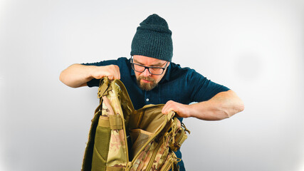 Portrait of handsome bearded man with beanie and glasses looking into camo military backpack,...