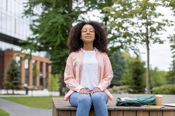 African American woman sitting outdoors in park enjoying moment of relaxation with closed eyes. Woman wearing casual attire enjoying tranquility in urban park setting. Coffee cup, backpack on bench.