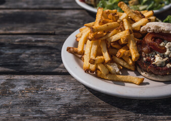 Hamburger with blue cheese, bacon and beef, accompanied by French fries, typical American food, served on a white plate on a rustic wooden table.