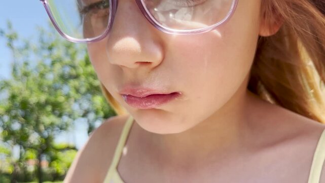 Close up on the face of an eight-year-old girl child eating ice cream with a spoon against a background of trees and a blue sky. A child eats ice cream with a spoon on the street.