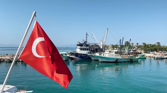 Beach tour boat departs harbor, a vibrant Turkish flag against a backdrop of docked fishing vessels and a serene coastal view. Flag of Turkey fluttering in breeze. &Ouml;ren Resort, Milas, Muğla, Turkey 
