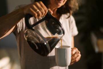 middle aged woman pouring coffee into mug on sunny morning