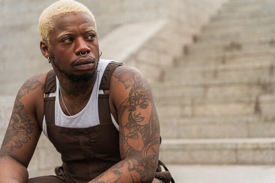 Young Stylish Man With Tattoos Sitting On Steps Looking Away