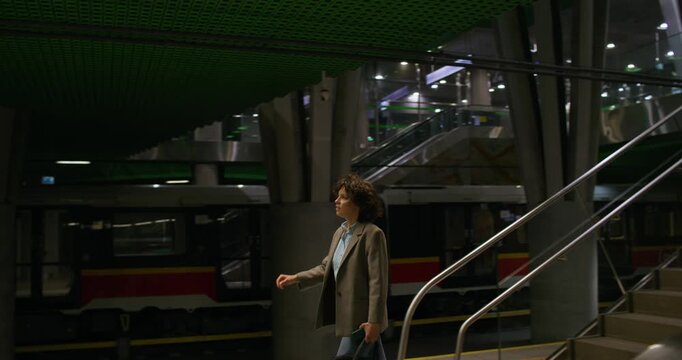 A stylish woman with a briefcase goes down to the railway platform. Dimmed lights. Metro station
