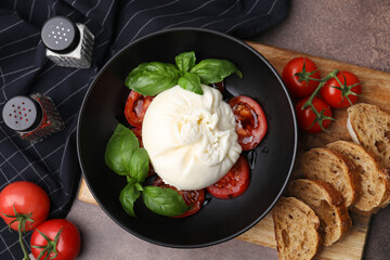 Delicious burrata cheese, tomatoes, basil and bread in bowl on brown table, flat lay