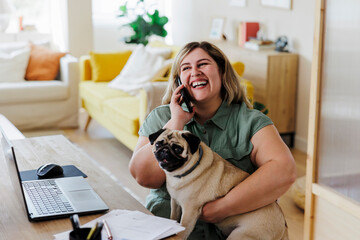 Happy woman hugging pug dog while talking on phone at home office