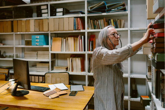 Senior woman in office looking for documents folder on bookshelf