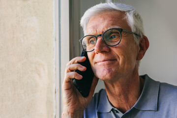 Senior man with glasses talking on smartphone by window, gray hair