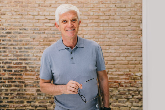 Senior man holding eyeglasses standing against brick wall background