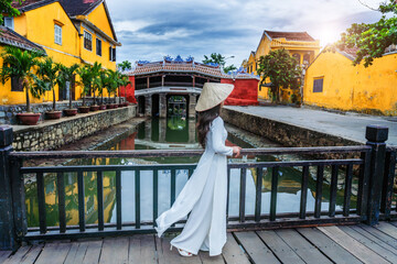 A young Vietnamese woman in an ao dai dress walks on a wooden bridge at the old town, hoi an, vietnam.