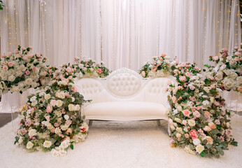 White sofa for the bride and groom decorated with flowers and garlands. Location for a wedding ceremony in oriental style. Beautiful white sofa with flowers for a traditional bride and groom ceremony