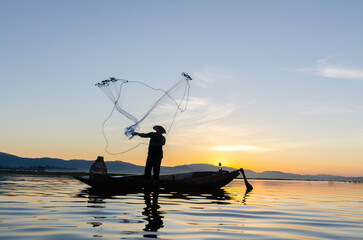 Asian fisherman on wooden boat casting a net for catching freshwater fish in nature river in the early morning before sunrise