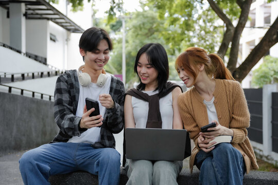 Group of three young asian people using technological devices and smiling while sitting on a bench outdoors