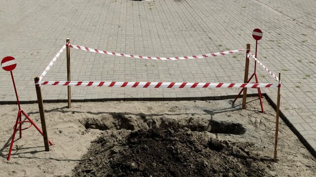 A construction site with a freshly dug hole is cordoned off with red and white tape, alongside warning signs, indicating the area is awaiting further work or inspection, emphasizing safety and caution