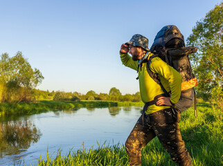 A hiker gazes thoughtfully across the tranquil river, surrounded by lush greenery and vibrant nature at golden hour.