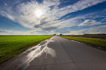 A tranquil landscape showcases an empty road stretching toward the horizon, flanked by green fields glistening from recent rain and bathed in warm sunlight.