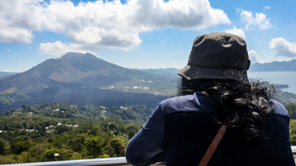 Back view of a female tourist standing on a viewpoint looking at Mount Batur. Volcanic landscape and surrounding greenery are visible under a clear sky, ideal for travel and adventure related content
