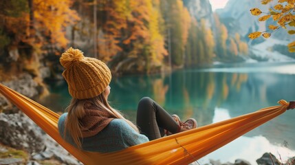 Rear view of woman in warm clothing relaxing on hammock among nature with lake, mountain and forest in Autumn time