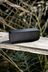 Black wireless bluetooth portable speaker on a wooden board in outdoor nature forest reeds...