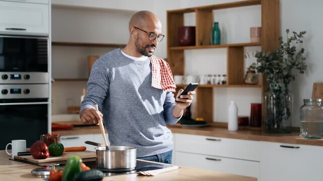 Video of handsome man cooking and using his mobile phone in the kitchen at home.