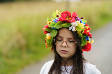 Naklejka premium A little girl in glasses and a national traditional Ukrainian wreath stands emotionally on a narrow rural road. Concept of national identity