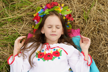 A little beautiful girl in a wreath and embroidered shirt lying on a freshly mown field. The...