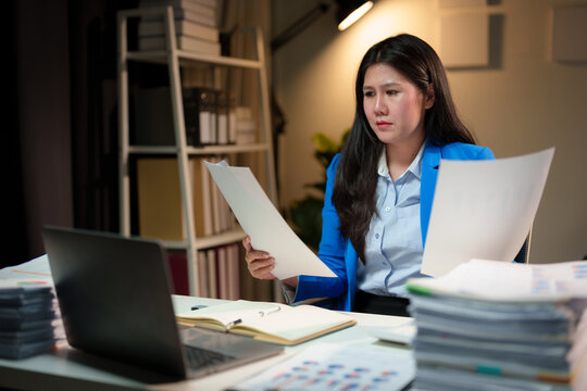 Stressed and tired Asian businesswoman feels a headache while working overtime at night with a pile of documents. Searching, sifting through documents, can't find them. Problems around the office.