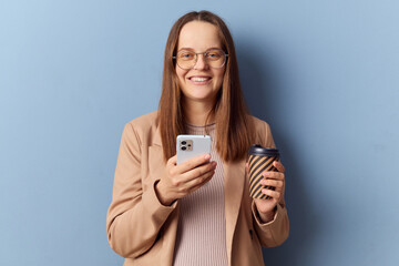Fototapeta premium Joyful delighted brown haired pregnant woman in glasses with big belly isolated over blue background scrolling through her smartphone having internet addiction enjoying online communication