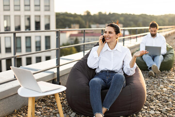 Business professionals working remotely on outdoor rooftop lounge, using laptops and smartphones. Relaxed work, casual attire, and modern workspace setting. Ideal for collaborative work scenes.